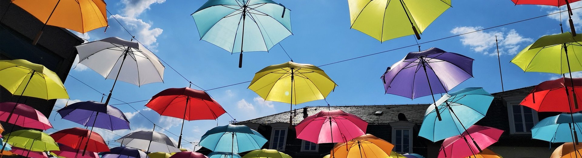 Viele bunte Regenschirme wurden hinter dem Rathaus Saarwellingen aufgehangen und bilden einen Schirmhimmel. Die Sonne scheint und der Himmel ist ist hellblau.