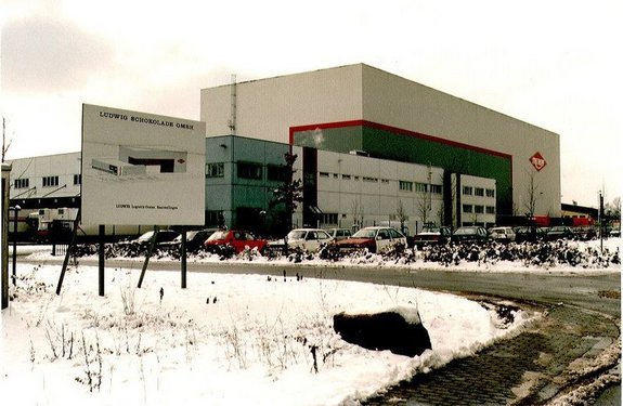 Das Bild zeigt ein großes Firmengebäude mit vorgelagertem Parkplatzbereich und Rasenflächen. Das Bild wurde bei Schnee aufgenommen. Am Gebäude ist ein rotes Logo, in welchem in weißer Schrift "Trumph" steht. Ein großes Hinweisschild vor dem Firmengelände weißt darauf hin, dass es sich bei der Firma um die Ludwig Schokoladen GmbH handelt.
