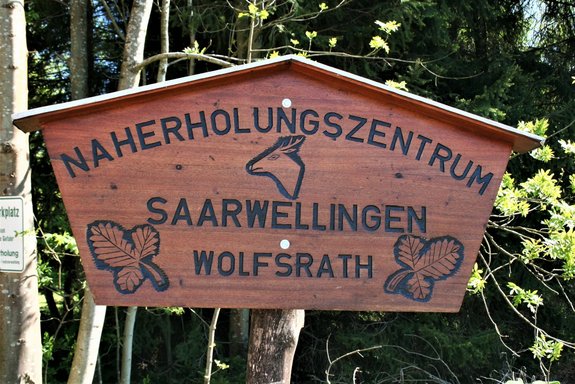 Das Bild zeigt ein Holzschild, worauf steht Naherholungszentrum Saarwellingen Wolfsrath. Zudem sind zwei Blätter und ein Tierkopf in das Holzschild geschnitzt.