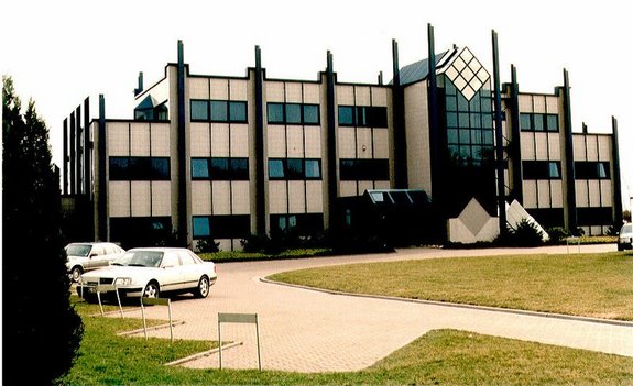 Das Bild zeigt ein großes Firmengebäude mit vorgelagertem Parkplatzbereich und Rasenflächen.