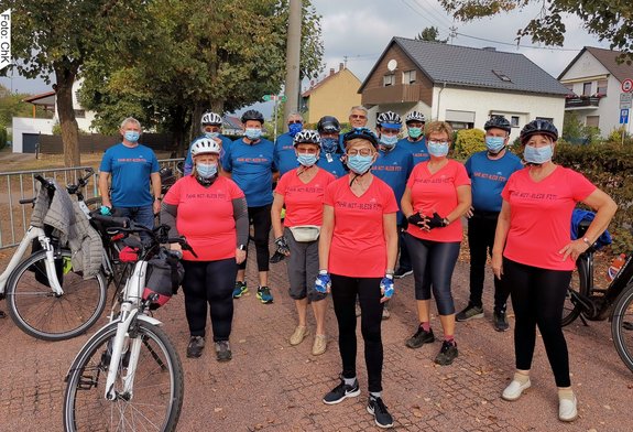 Personen mit blau- und rotfarbenen T-Shirts. Auf den T-Shirts steht: Fahr mit - Bleib fit. Die Personen tragen Mundschutz und Helme. Links unten im Bild stehen zwei weiße Fahrräder.