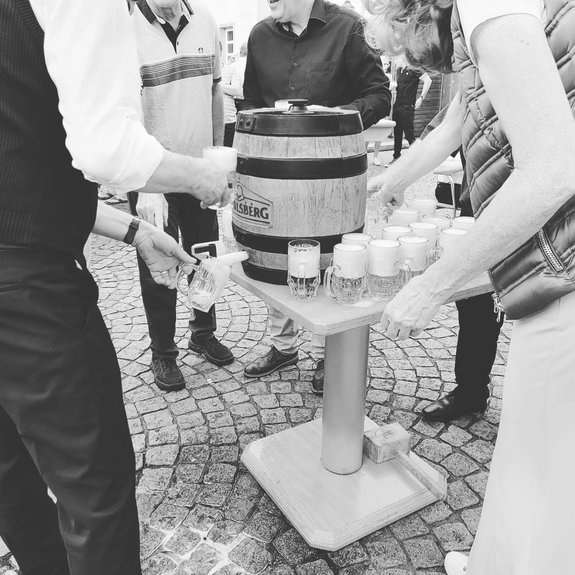 Schwarzweißes Foto, welches den traditionellen Fassanstich beim Saarwellinger Schlossfest zeigt. Ein kleiner Tisch in der Bildmitte, auf dem ein Bierfass und einige Biergläser stehen. Um den Tisch stehen mehrere Personen, welche dabei sind, Bier zu zapfen.