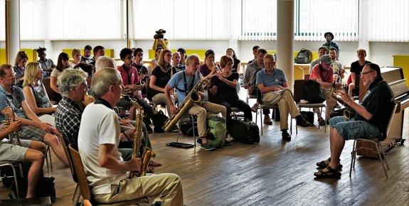 Gruppe von Musiker*innen sitzen im Halbkreis vor einem Musiklehrer