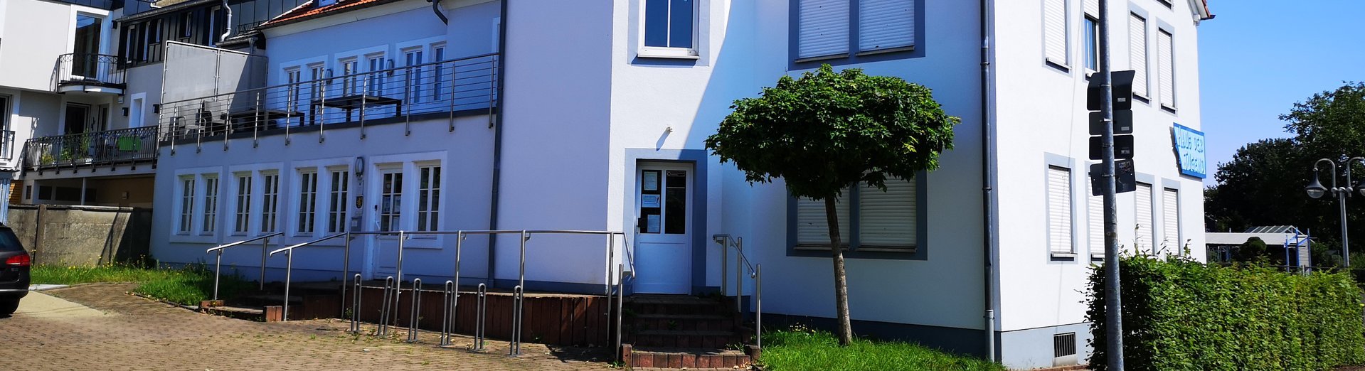 Das Bild zeigt die Rückansicht vom Haus der Jugend in der Engelstraße in Saarwellingen. Es ist ein hellblaues, dreistöckiges Haus. Der Eingang zum Amt für Jugend, Senioren und Soziales befindet sich hinter dem Haus und ist somit auf dem Foto sichtbar.