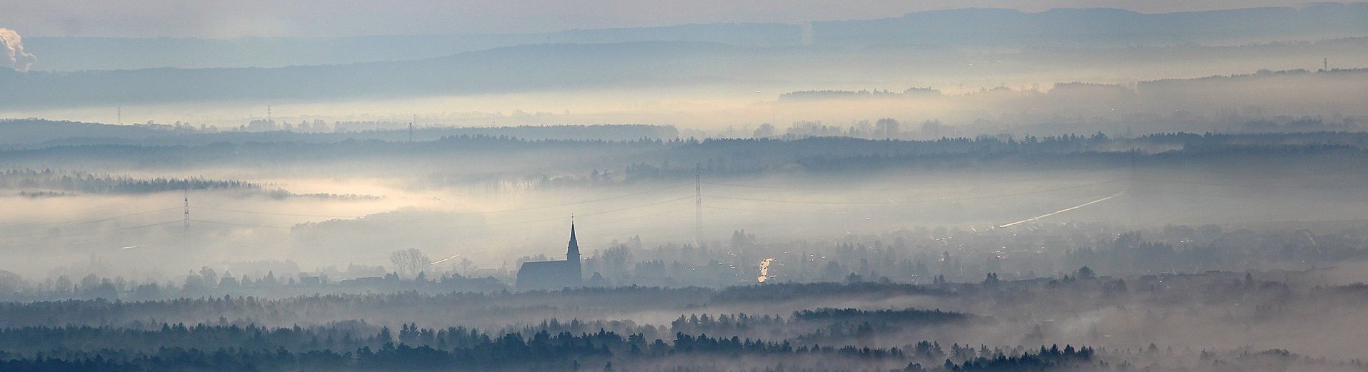 Blick über eine Landschaft, welche teilweise von Nebel überdeckt ist. Eine Kirchenspitze ragt aus dem Nebel.