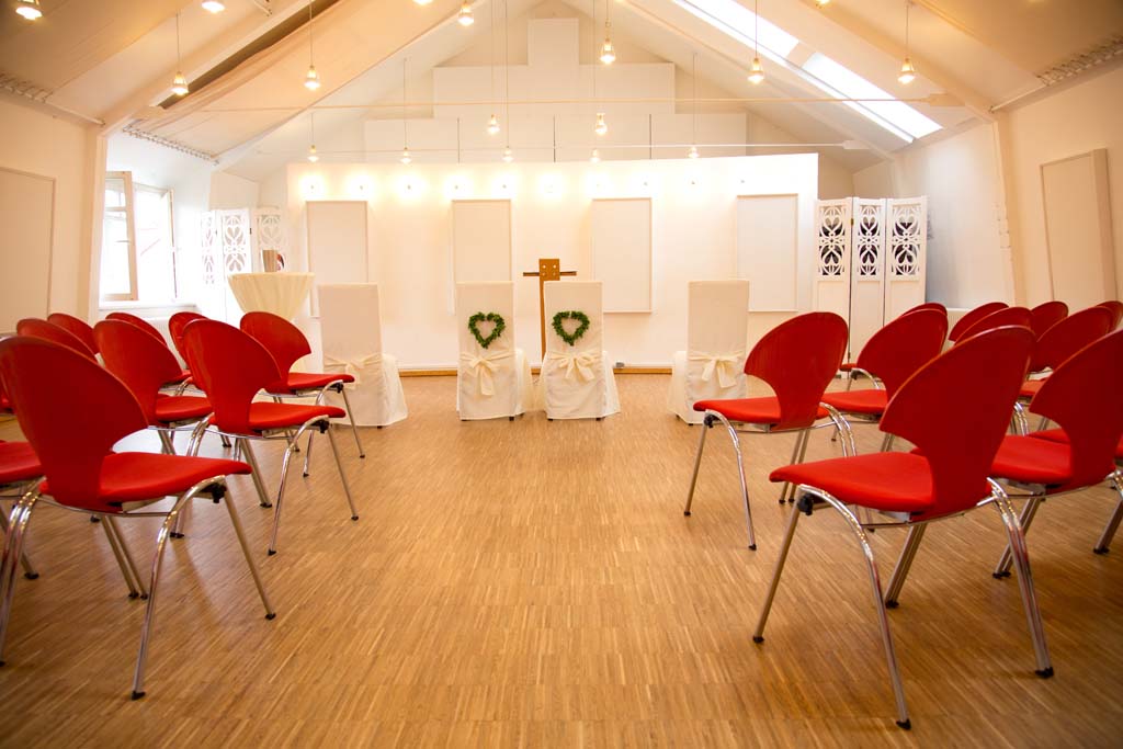 Blick in den Festsaal "Altes Rathaus". Holzboden mit Stäbchenpakett, Spitzdecke mit hellen Akustikvorhängen vor den Fenstern und weiße Wände. Der Raum ist für eine Trauung vorbereitet. Rote Stuhlreihen stehen hinter dem Traubereich. Der Traubereich besteht aus vier Stühlen mit Weißen Hussen. Die Brautpaarstühle sind an der Rückenlehne zudem je mit einem Herz aus Blumen geschmückt.
