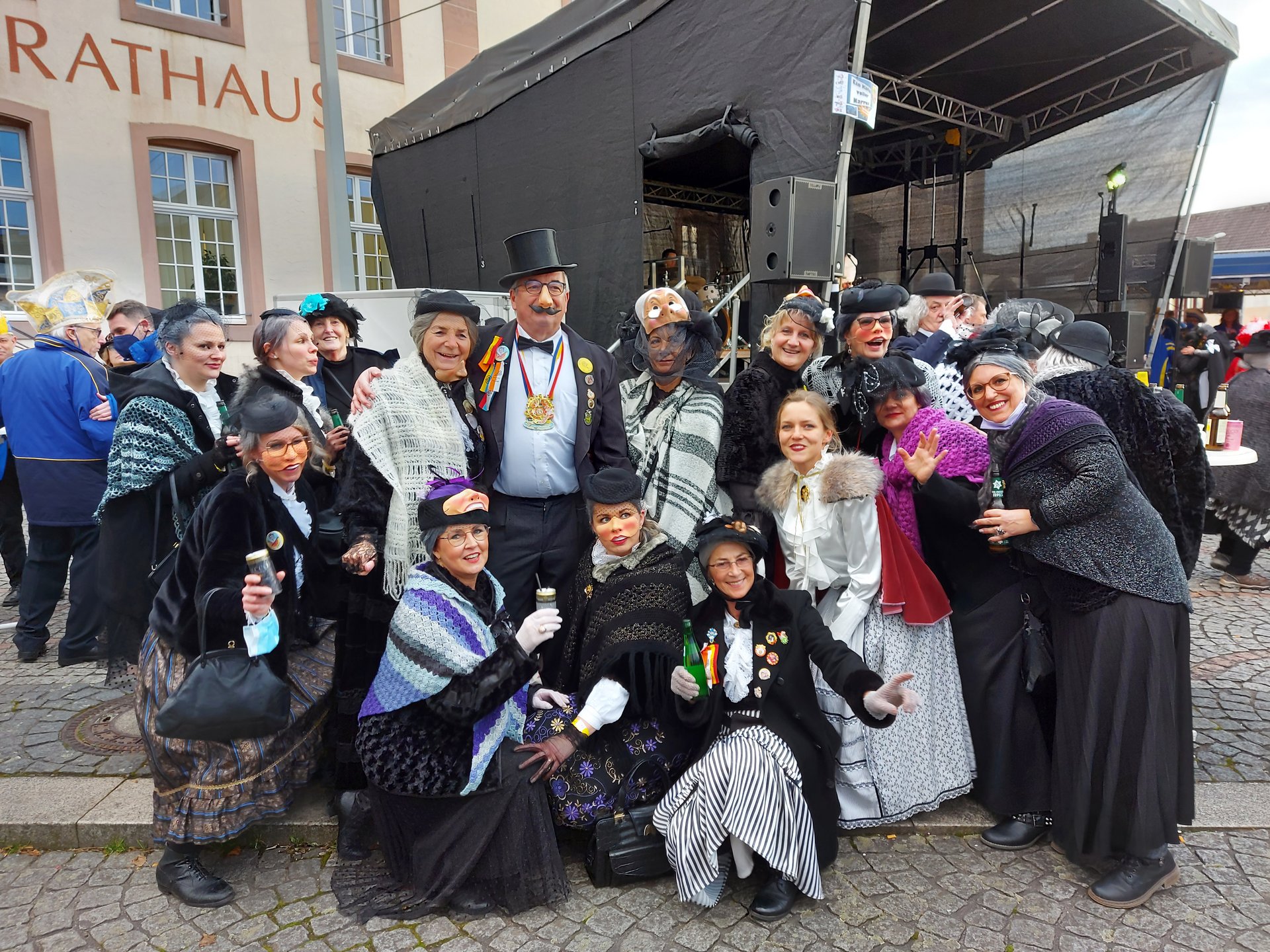 Vor dem Rathaus steht und kniet eine Gruppe verkleideter Menschen und schaut in die Kamera. Im Hintergrund erkennt man eine Bühne und einige weitere Personen. Das Bild zeigt sogenannte Greesen am Saarwellinger Greesentag.