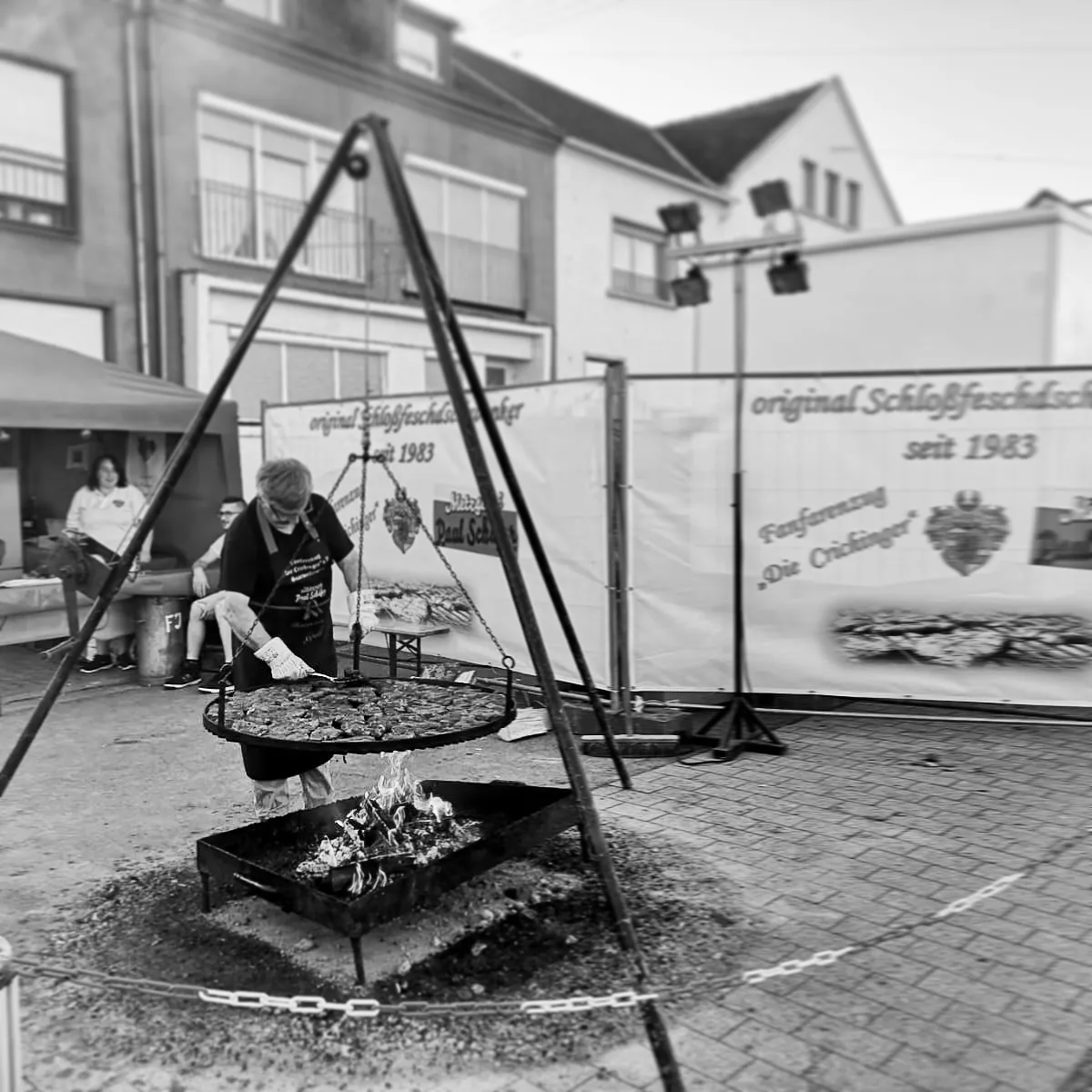 Schwarzweißes Foto. Es zeigt einen Essensstand am Saarwellinger Schlossfest, an dem es Schwenkbraten vom saarländischen Schwenker gibt. Ein Mann steht an dem großen, runden Schwenkrost, welcher an einem dreibeinigen Metallgerüst befestigt ist. Darunter brennt das Feuer.