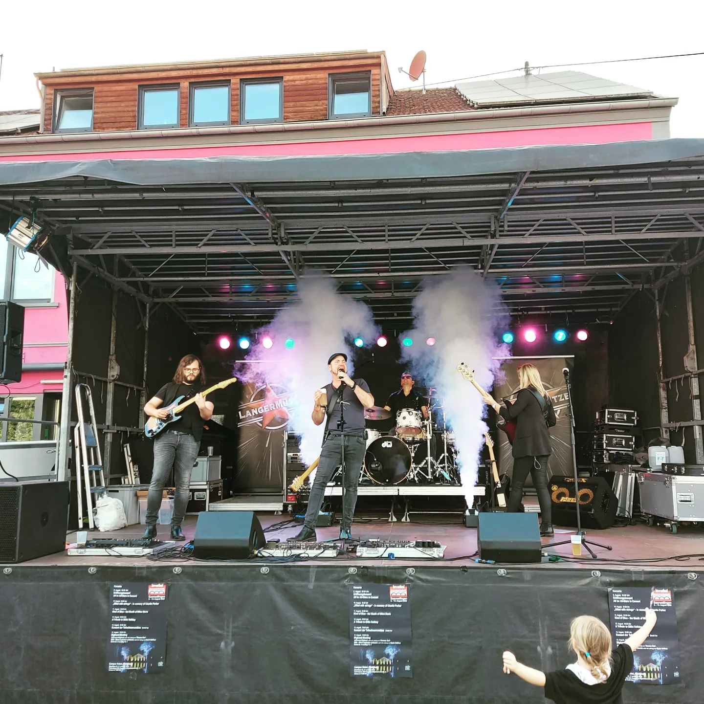 Blick auf die Blumen-Schäfer-Bühne beim Saarwellinger Schlossfest. Ein kleines Kind stehr jubelnd davor. Auf der Bühne sieht man eine vierköpfige Band, welche Musik macht. Die Bühne ist in verschiedenen Farben beleuchtet und es steigt Effektnebel auf.