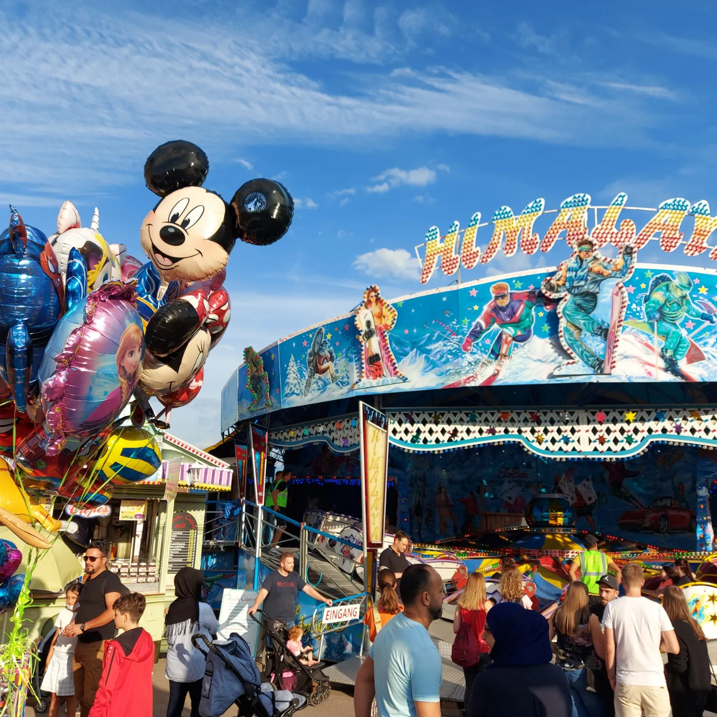 Blick auf die Sommerkirmes beim Saarwellinger Schloßfest. Blauer, fast wolkenloser Himmel. Bunte Heliumballons auf der linken Bildseite. Im Hintergrund die bunte Berg- und Talbahn "Himalaya", wovor einige Menschen stehen.