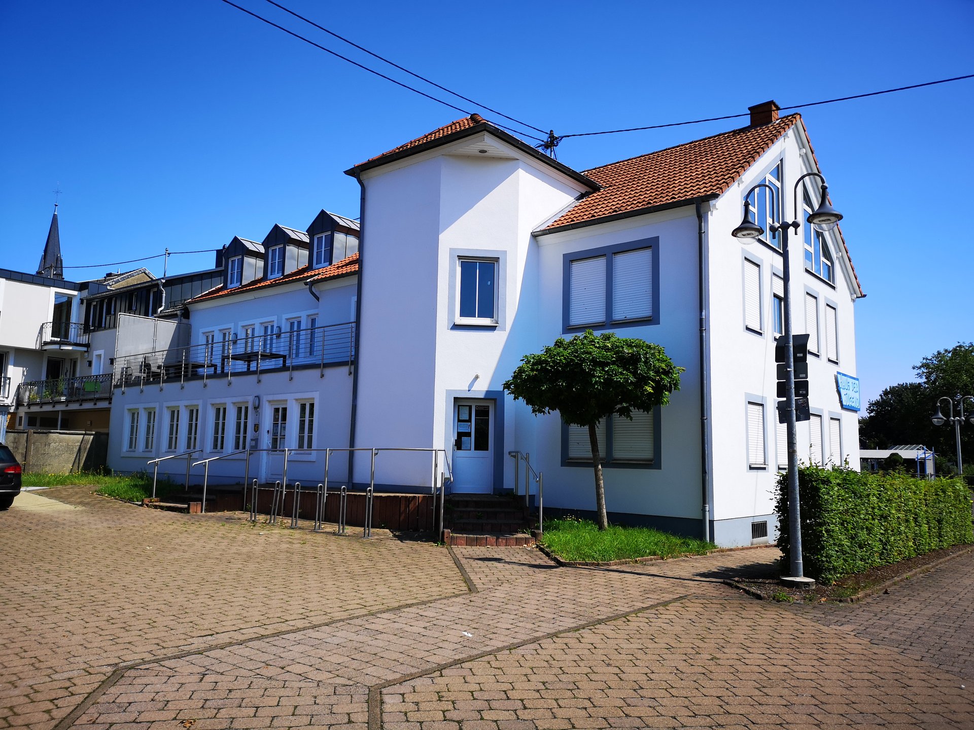 Das Bild zeigt die Rückansicht vom Haus der Jugend in der Engelstraße in Saarwellingen. Es ist ein hellblaues, dreistöckiges Haus. Der Eingang zum Amt für Jugend, Senioren und Soziales befindet sich hinter dem Haus und ist somit auf dem Foto sichtbar.